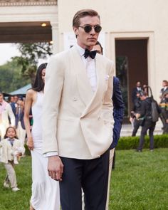 a man in a tuxedo and sunglasses standing on the grass with people behind him