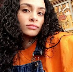 a woman with long black curly hair wearing an orange shirt and overalls is looking at the camera