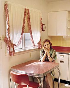 a woman sitting at a kitchen table in front of a window with red and white curtains