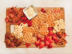 waffles and strawberries are arranged on a cutting board with bacon, banana slices, and butter