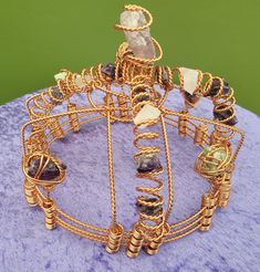 a gold wire sculpture with many different beads and stones on it's sides, sitting on a purple surface