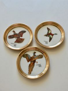 three plates with birds painted on them are sitting on a table top, one is gold and the other is white