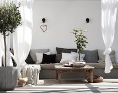 an outdoor living room with white curtains and pillows on the couch, potted plants