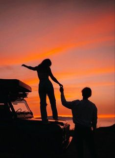 a man holding the hand of a woman while standing on top of a car at sunset