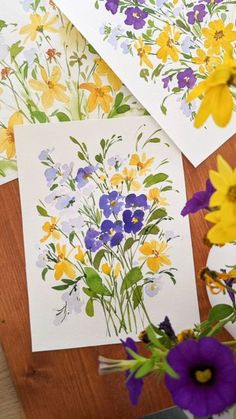 three cards with flowers on them sitting next to a vase filled with purple and yellow flowers