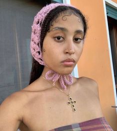 a young woman wearing a pink crochet headband and a cross necklace, standing in front of a window