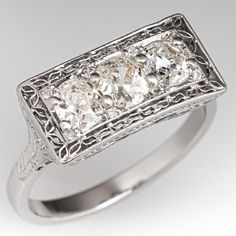 This Art Deco circa 1920s diamond ring features a pierced design with engraved details and bordered with milgrain edging. The ring is centered with three (3), shared prong set, old mine cut diamonds and bordered with eight (8), bead set, round single cut diamonds. The shoulders of the ring are each accented with one (1), bead set, round single cut diamonds. The ring measures 8.8mm at the top, rises 4.9mm above the finger, tapering to 2.3mm wide and 1.4mm thick at the base of the shank. It is cur Antique Three-stone Diamond Rings, Antique Three Stone Diamond Ring In Diamond White, Antique Oval Three Stone Diamond Ring, Antique Platinum Three Stone Diamond Ring, Antique Platinum Three Stone Rings, Antique Three Stone Platinum Rings, Antique Three Stone Diamond Ring For Formal Occasions, Antique Platinum Three-stone Jewelry, Heirloom Three Stone Platinum Ring