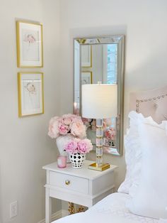 a white nightstand with pink flowers on top and a mirror above it next to a bed