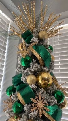 a christmas tree with green and gold ornaments on it's top, sitting in front of a window