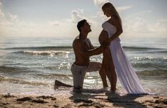 a man kneeling down next to a pregnant woman on the beach in front of the ocean