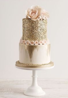 a three tiered cake with gold and white frosting, topped with a pink flower