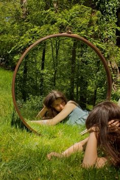 two women laying on the grass in front of a circular mirror with trees behind them