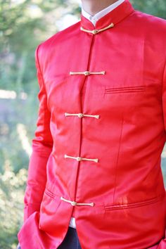 a man wearing a red jacket with gold buttons on the chest and sleeves, standing in front of some trees