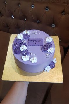 a purple cake with white flowers on it sitting on a wooden tray next to a person's hand