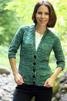 a woman wearing a green knitted cardigan standing in front of rocks and trees