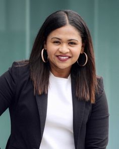 a woman with large hoop earrings standing in front of a green wall wearing a black blazer and white top