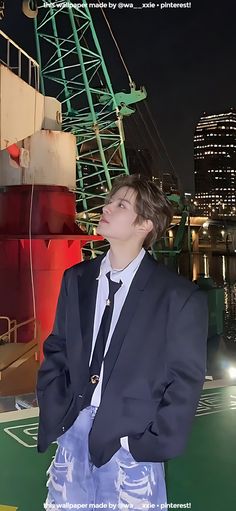 a man in a suit and tie standing next to a boat at night with the city lights behind him