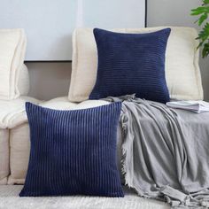 a couch with pillows and blankets on it in front of a white sofa, next to a potted plant