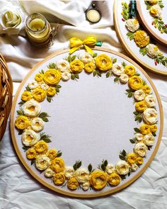 three yellow roses are arranged in the center of two embroidery hoops on a white tablecloth