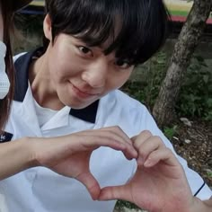 two people making a heart shape with their hands while standing next to each other in front of trees