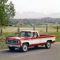 a red and white pickup truck driving down the road in front of a farm house