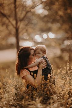 a woman holding a baby in her arms while standing in tall grass with trees behind her