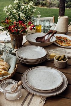 an outdoor table with plates and bowls on it, flowers in vases next to them