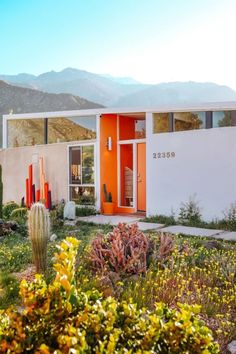 The photo shows the outside of the front of the house. It has a bright orange door with a window to the left of it that looks into the front entryway. The house is white with bright orange around the door area and it slants to the right side. There is a large window on the left side of the house and windows all along the top of the house. The front yard has several cactus plants, as well as yellow and purple flowers. There are mountains in the background behind the house. William Krisel, Mid Century Modern House Exterior, Mid Century Modern Exterior, Eclectic Homes, Palm Springs Home, Mid Century Exterior, 70s House, Desert House, Dwell Magazine