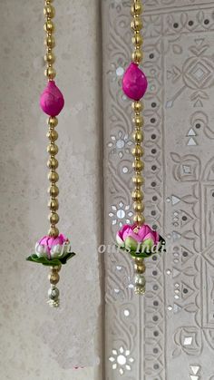 two pink flowers hanging from gold chains on a wall next to a vase with white flowers in it