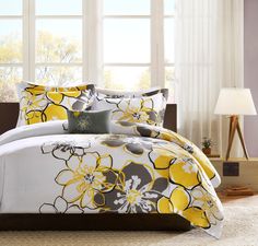 a bed with yellow and gray flowers on it in front of a large window next to a lamp