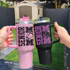 two women holding up pink and black tumblers in front of a fenced yard