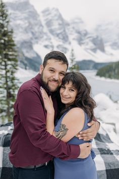 a man and woman hugging each other in front of snow covered mountains with trees on either side