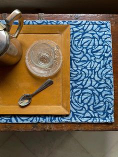 a tea pot, spoon and glass on a wooden tray with a blue patterned place mat