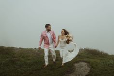 a bride and groom are walking in the fog together on top of a grassy hill