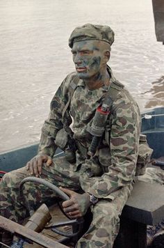 a man in camouflage sitting on the back of a boat with his face painted like a soldier