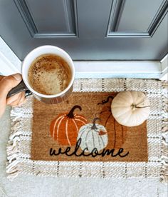 someone holding a cup of coffee in front of a welcome mat with pumpkins on it