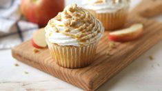 two cupcakes with white frosting and apple slices on a cutting board next to them