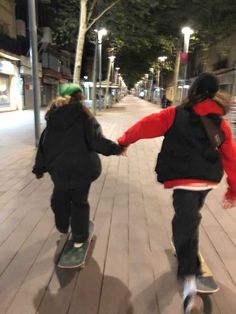 two people riding skateboards down a sidewalk next to a street at night with one holding the other's hand