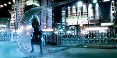 a woman is walking down the street in front of some tall buildings at night time