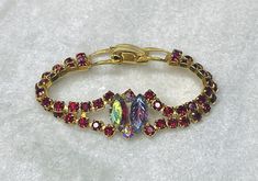 A 50's colorful bracelet of red rhinestones in a festoon design, faced with two aurora borealis baguette stones, one faceted and the other pressed in a leaf pattern. It closes with a hinged clip,  It measures 7" long and 3/8" wide. The face is 3/4". Excellent condition. Catalog # 8844 Red Jubilee Bracelets For Parties, Red Jubilee Bracelet For Party, Vintage Red Bracelets For Party, Red Jeweled Bracelet For Party, Adjustable Red Bracelet With Rhinestones, Red Vintage Party Bracelets, Formal Red Crystal Bracelets, Retro Red Bracelet Jewelry, Red Crystal Rhinestone Bracelet For Party