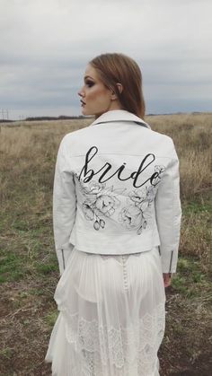 a woman standing in a field wearing a white jacket with the word bride written on it