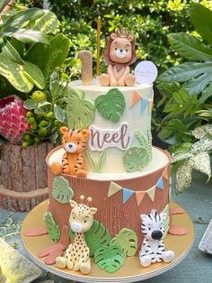 a three tiered cake decorated with jungle animals and trees is sitting on a table surrounded by greenery