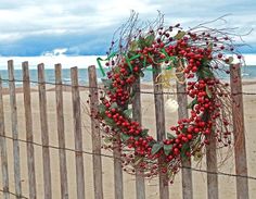 a wreath is hanging on a fence at the beach with red berries and green leaves