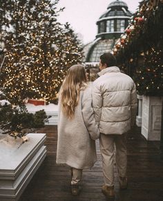 a man and woman walking down a wooden walkway in front of a christmas tree covered with lights