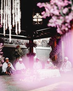 several people sitting at a table with flowers in front of them and a chandelier hanging from the ceiling