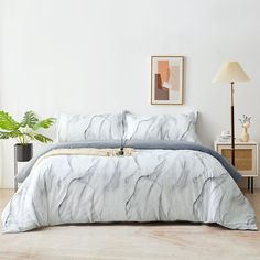 a bed with white and grey sheets in a room next to a plant on the floor