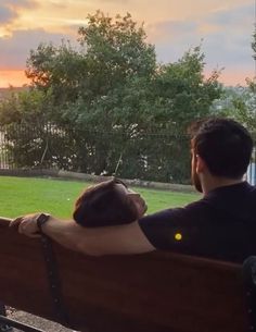 a man and woman sitting on a bench looking at the sunset over a grassy field