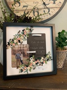 a wedding photo frame with flowers and greenery in front of a wooden wall clock