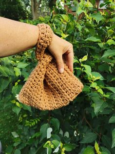 a hand is holding up a crocheted item in front of some bushes and trees