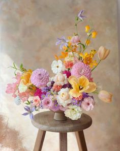 an arrangement of flowers on a stool in front of a wall with a window behind it